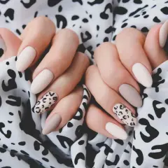 Dłonie z eleganckim panterkowym manicure w odcieniach beżu i ecru na tle wzorzystej tkaniny.