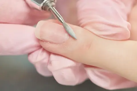 Zbliżenie na paznokieć, na którym frezarka usuwa odrost hybrydy. Dłonie w różowych rękawiczkach.