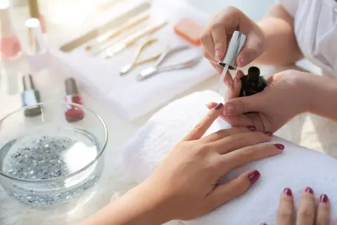 Dłoń podczas malowania paznokci ciemnym lakierem, w tle przybory do manicure.