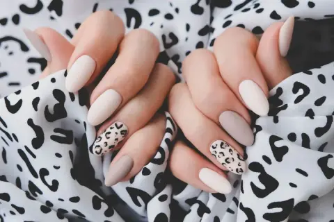 Dłonie z eleganckim panterkowym manicure w odcieniach beżu i ecru na tle wzorzystej tkaniny.