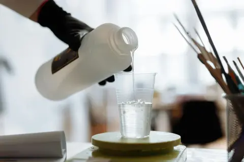 Ręka w czarnej rękawiczce przelewa przezroczysty płyn z butelki do plastikowego kubka stojącego na wadze.
