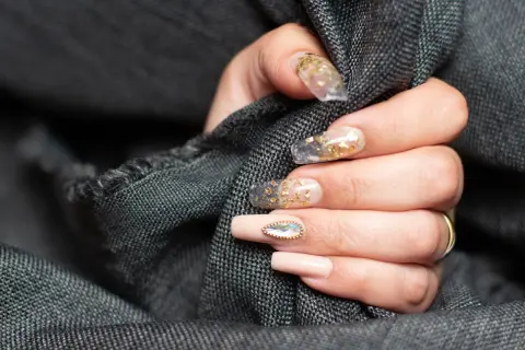Dłoń z paznokciami w stylu szklanego manicure z płatkami złota i ozdobnym kamieniem.