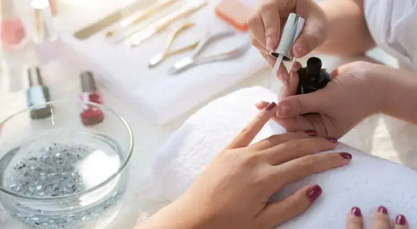 Dłoń podczas malowania paznokci ciemnym lakierem, w tle przybory do manicure.