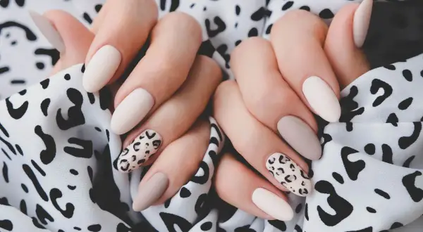 Dłonie z eleganckim panterkowym manicure w odcieniach beżu i ecru na tle wzorzystej tkaniny.