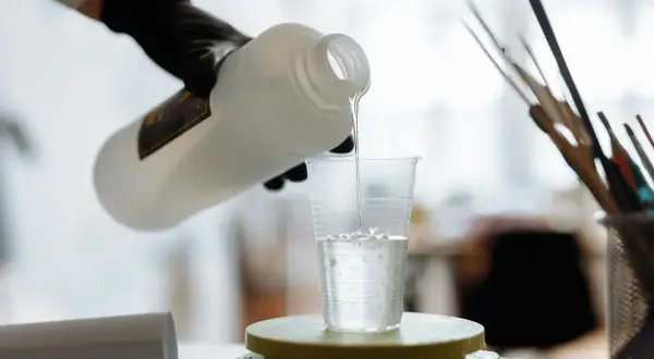 Ręka w czarnej rękawiczce przelewa przezroczysty płyn z butelki do plastikowego kubka stojącego na wadze.