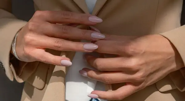 Dłonie kobiety z delikatnym, jasnoróżowym manicurem w beżowej marynarce.