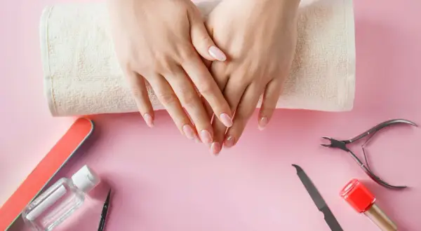 Dłonie z paznokciami na ręczniku, otoczone narzędziami do manicure: pilnik, nożyczki, cążki, odżywka i lakier.