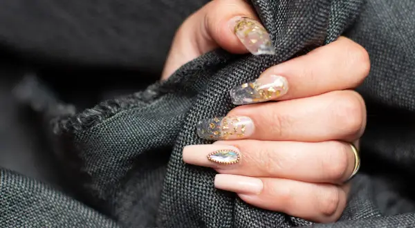 Dłoń z paznokciami w stylu szklanego manicure z płatkami złota i ozdobnym kamieniem.