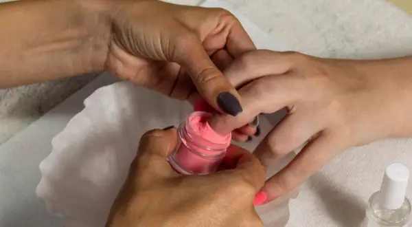 Dłoń zanurza paznokieć w różowym pudrze do manicure tytanowego.