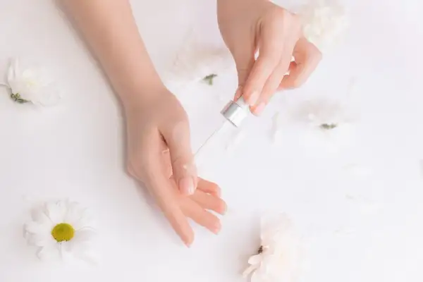 Dłonie z pipetką aplikującą olejek na paznokieć, otoczone białymi kwiatami.