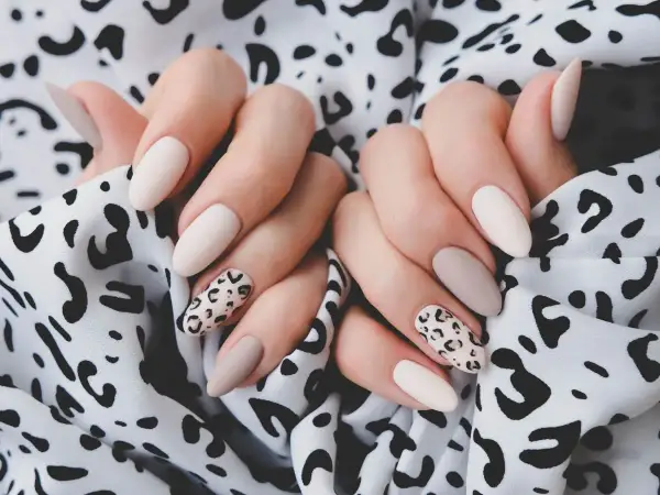 Dłonie z eleganckim panterkowym manicure w odcieniach beżu i ecru na tle wzorzystej tkaniny.