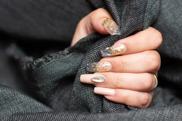 Dłoń z paznokciami w stylu szklanego manicure z płatkami złota i ozdobnym kamieniem.